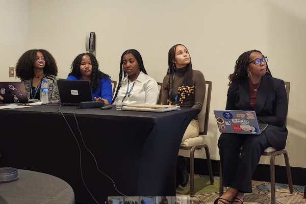 A group of women is gathered around a table, each with their laptops open.