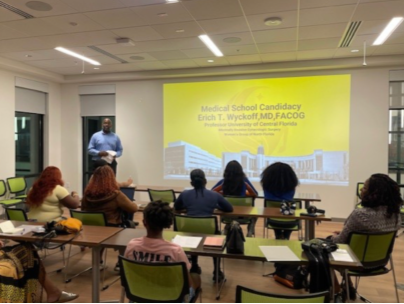 A classroom setting where a person is standing in front of a projection screen, with students sitting and paying attention.